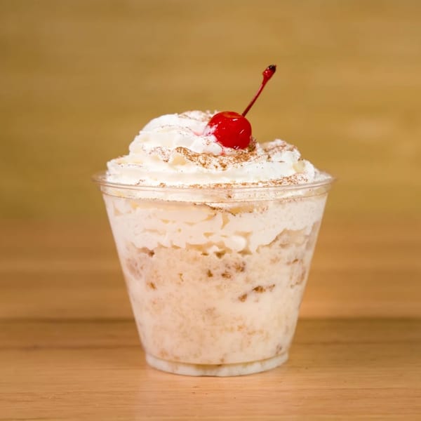 A clear plastic cup filled with a creamy dessert, topped with whipped cream, cinnamon, and a cherry.