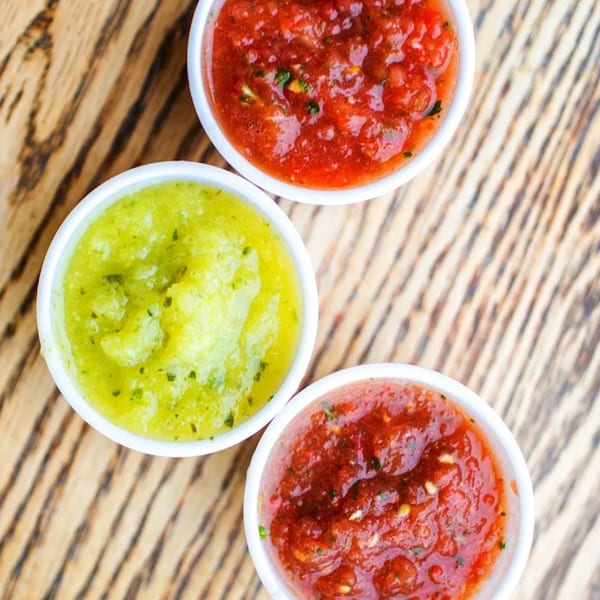 Three small white bowls of Don Juan Mex Grill salsa, two red and one green, on a wooden table.