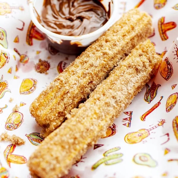 Two sugar-dusted churros with a side of chocolate dipping sauce on a festive Mexican-themed paper.