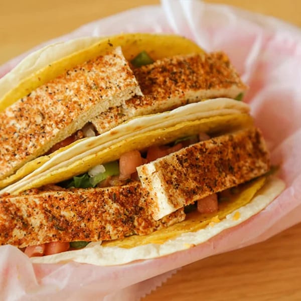 Two Don Juan Mex Grill tacos with seasoned tofu, lettuce, and tomatoes in a pink paper.