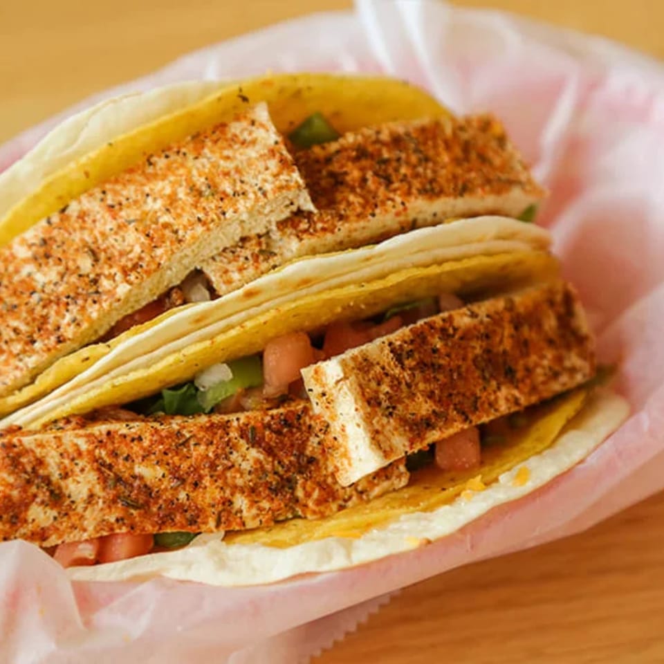 Two Don Juan Mex Grill tacos with seasoned tofu, lettuce, and tomatoes in a pink paper.