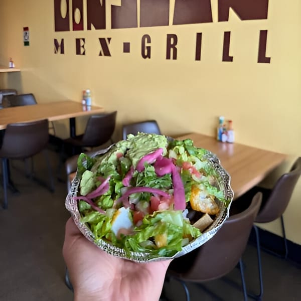 A hand holds a Don Juan Mex Grill burrito bowl with lettuce, guacamole, and pickled onions.