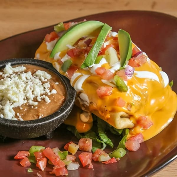 A Don Juan Mex Grill burrito covered in cheese sauce, sour cream, avocado, and pico de gallo, with a side of refried beans.
