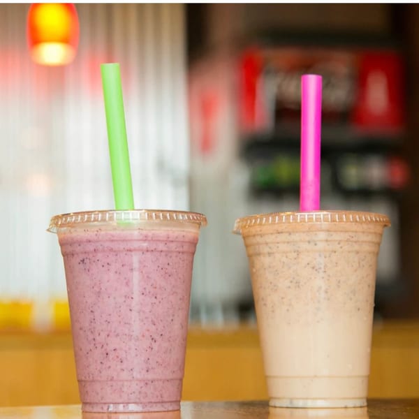 Two smoothies, one purple with a green straw and one tan with a pink straw, on a counter.