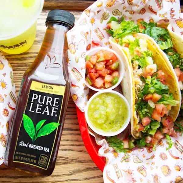Two Don Juan Mex Grill tacos with salsa, guacamole, and a Pure Leaf iced tea on a wooden table.