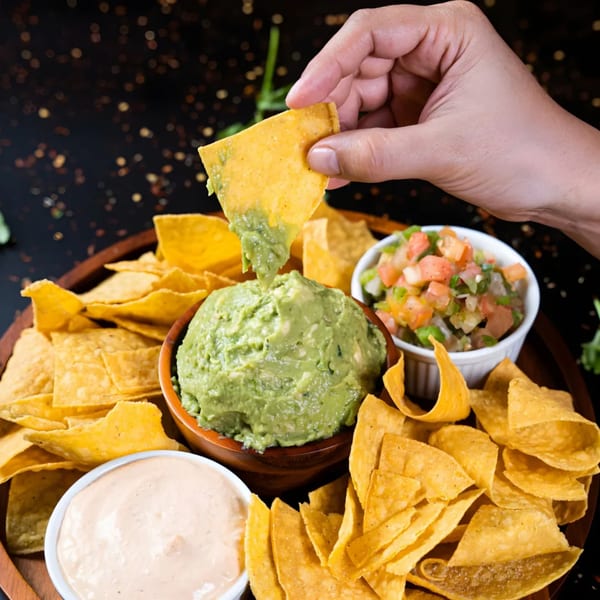 A hand dips a tortilla chip into a bowl of guacamole, surrounded by salsa and queso.