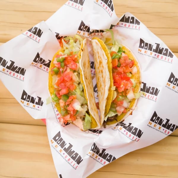 Two Don Juan Mex Grill tacos with lettuce, diced tomatoes, and onions in branded paper.